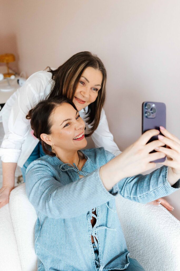Kaksi naista hymyilee ja poseeraa selfiessä valoisassa, kodikkaassa huoneessa. Toinen istuu ja pitää puhelinta, toinen seisoo takana. Kuva on ST Creative Studion kotisivuilla.